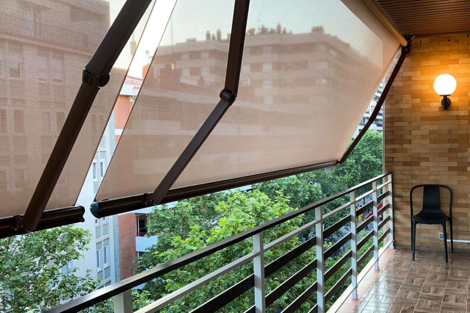 Balcón con toldos color canela, barandilla de metal, pared de ladrillo, silla negra y árboles.