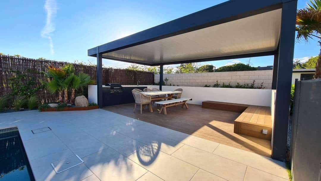 Patio exterior con pérgola oscura, juego de comedor y banco incorporado, junto a una piscina.