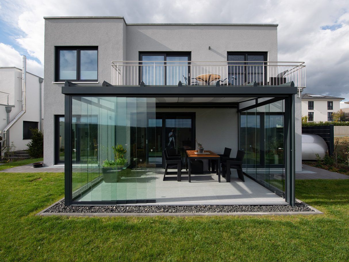 Casa moderna con patio acristalado y balcón superior; exterior gris, césped verde. Mesa y sillas visibles.