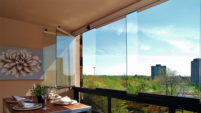 Balcón con paneles de vidrio, mesa de comedor y vista a la ciudad. Día soleado con cielo azul.