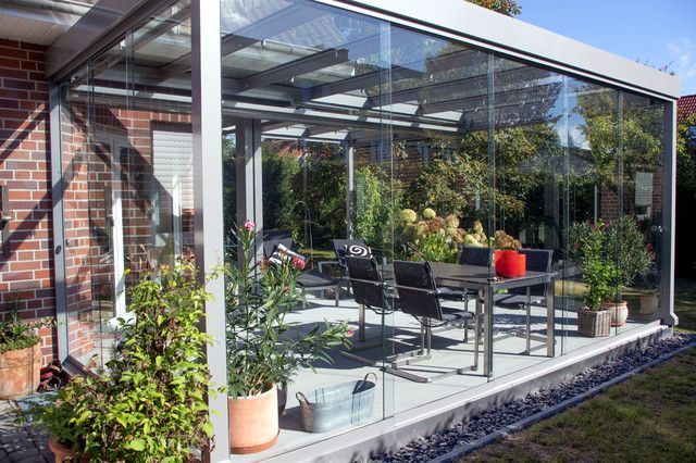 Patio cerrado con vidrio, mesa y sillas, plantas en macetas y casa de ladrillo.