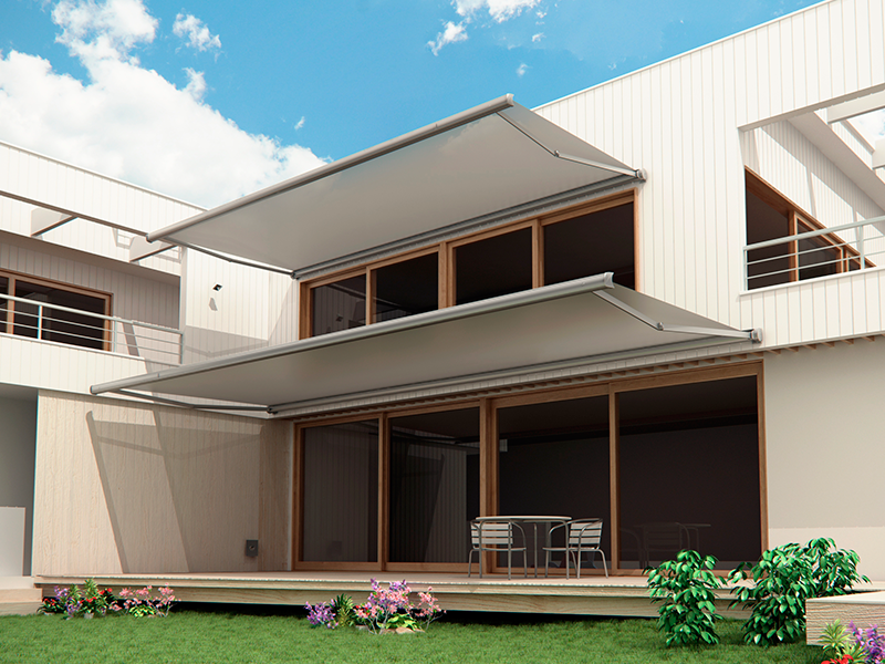 Casa moderna con dos toldos extensibles sobre grandes ventanales. Terraza de madera, césped verde y cielo azul.