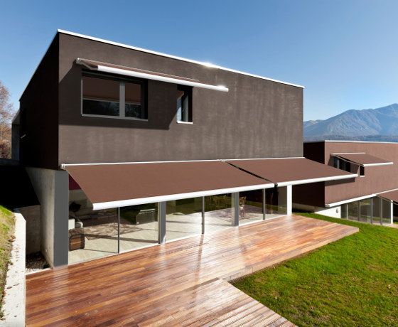 Casa moderna con toldos marrones sobre puertas de vidrio, terraza de madera y césped.