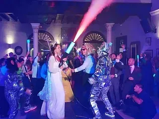 Una novia y un novio están bailando en una fiesta con un hombre con un traje de espejo que sostiene un cañón de confeti.