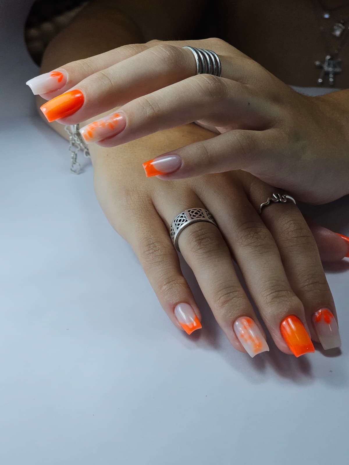 Manos con uñas con puntas naranjas, con anillos, apoyadas sobre una superficie blanca.