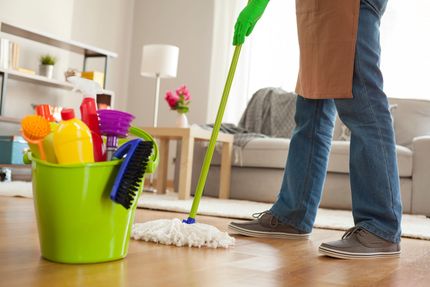 Eine Person putzt ein Wohnzimmer mit einem Wischmopp und einem Eimer voller Reinigungsmittel.