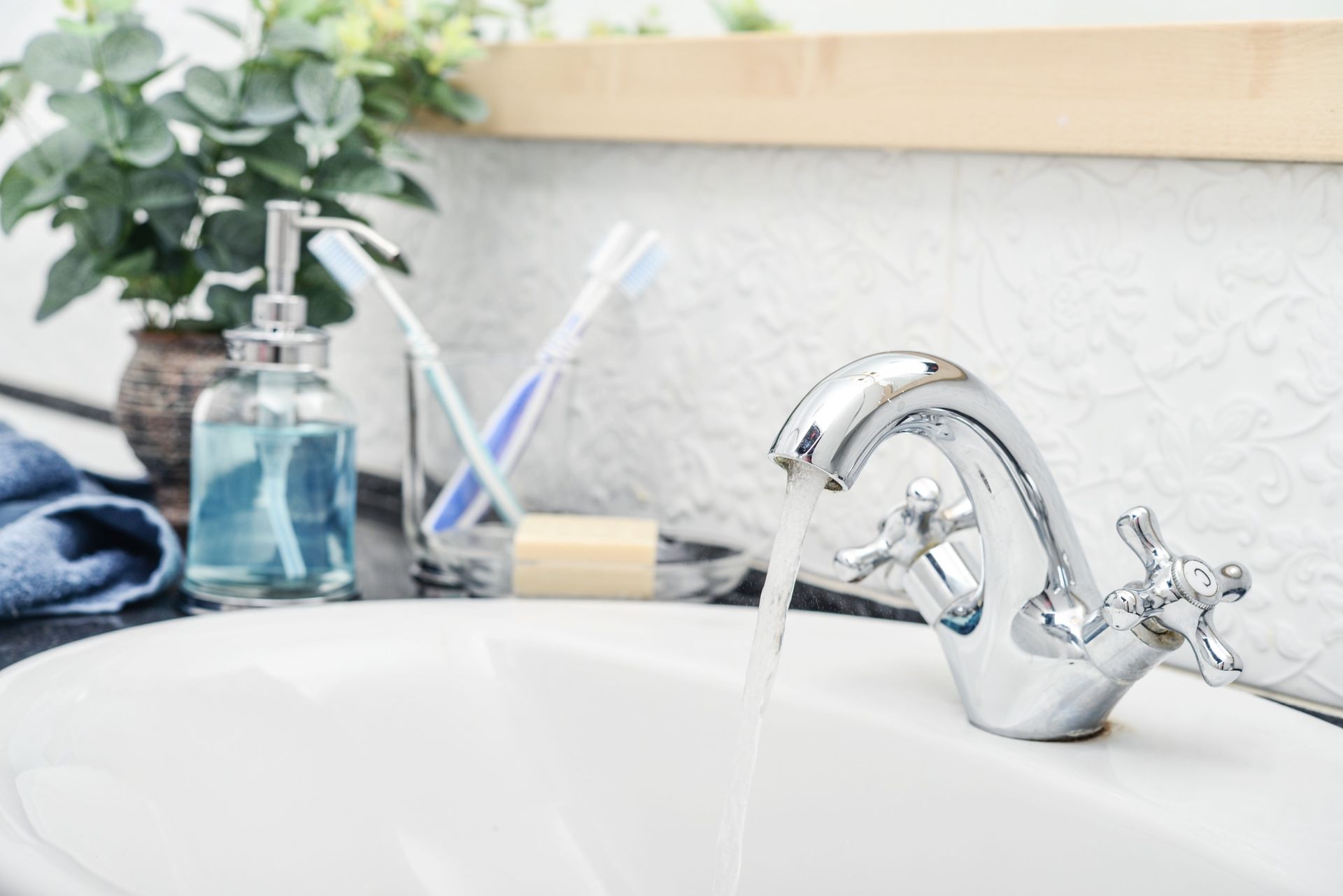 Un grifo cromado con agua corriente sobre un lavabo blanco en un baño. Al fondo se ven un dispensador de jabón, cepillos de dientes y una planta.