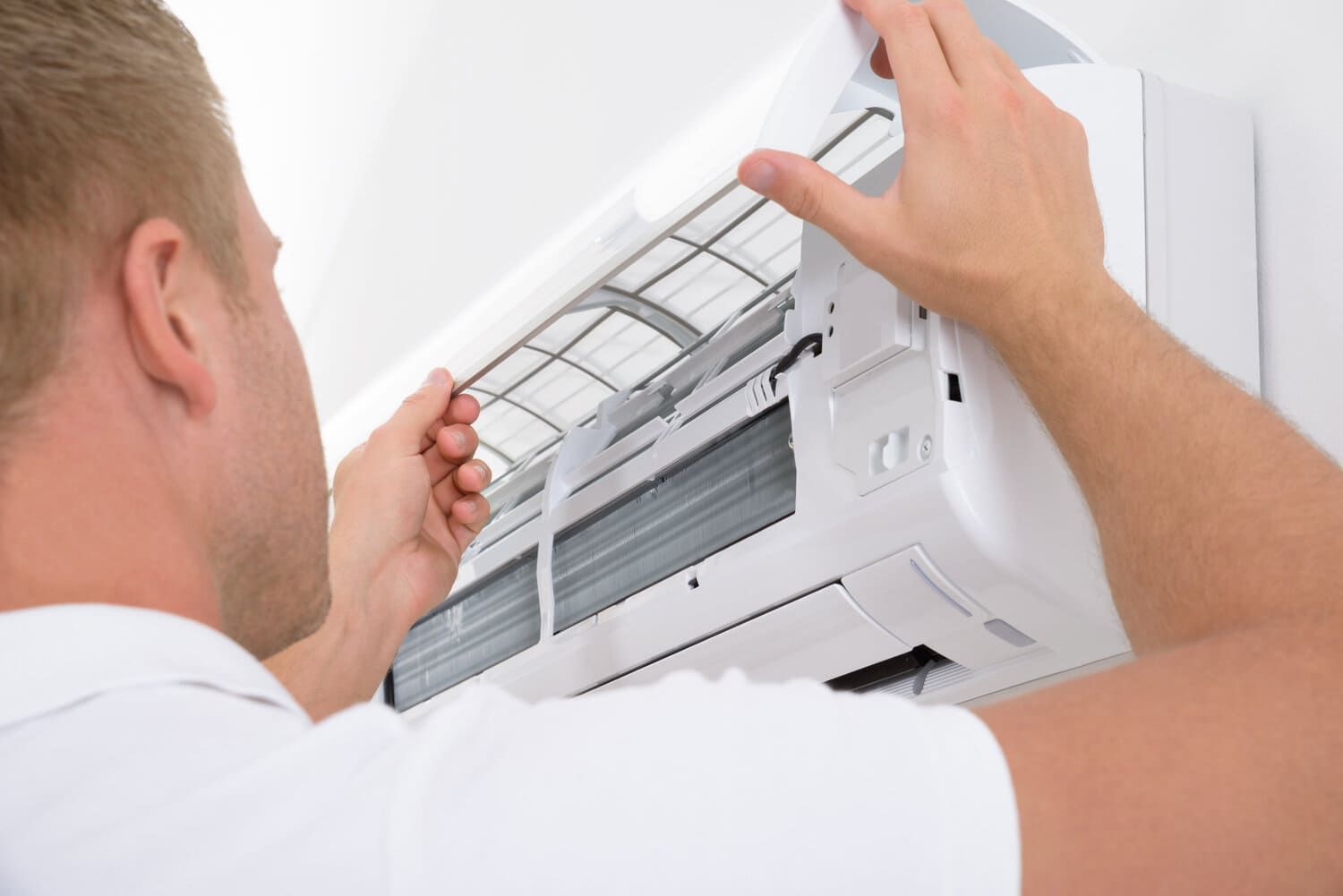 Hombre retirando un filtro de aire de un aire acondicionado de pared. Lleva una camisa blanca, con el aparato y la pared blancos.