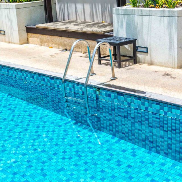 Piscina con paredes de azulejos azules, una escalera de metal y un taburete negro en el borde de hormigón.