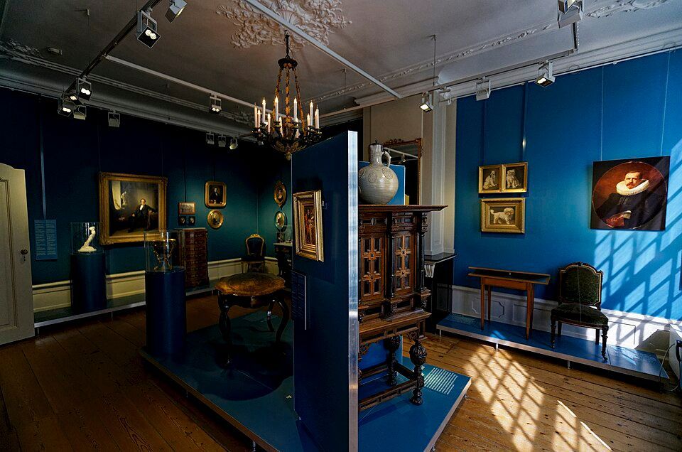 Blue-walled art gallery with framed paintings, wooden furniture, and sunlight casting window shadows on the floor