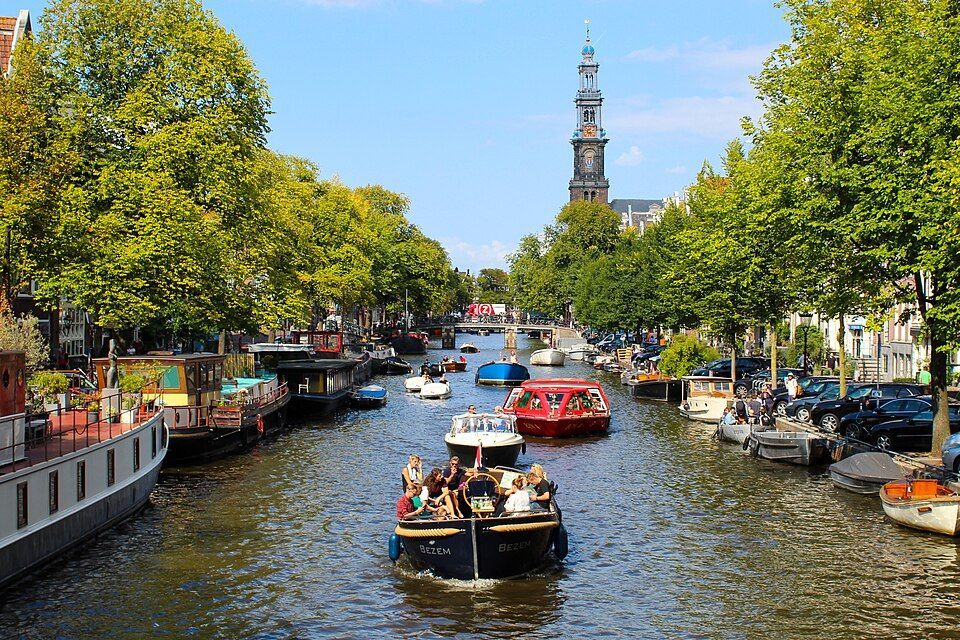 Boats on a tree-lined canal in a city, with a tall church tower in the distance under a blue sky