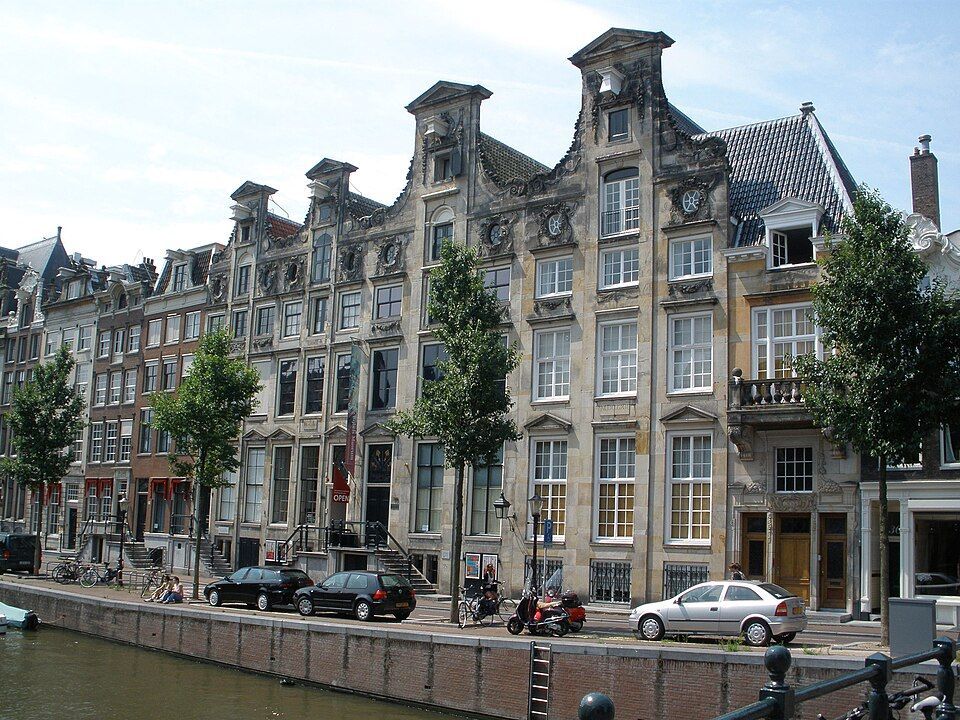 Canal-side row of ornate historic townhouses with parked cars and trees under a bright sky
