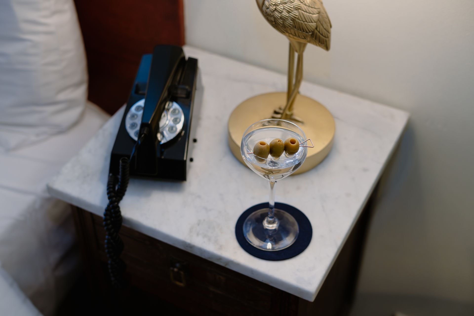 A bedside table with a black telephone, gold lamp, and a wine glass on a coaster