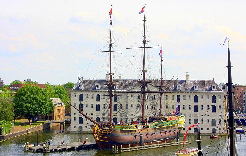 Historic sailing ship docked in front of a large white waterfront building with trees and a canal.