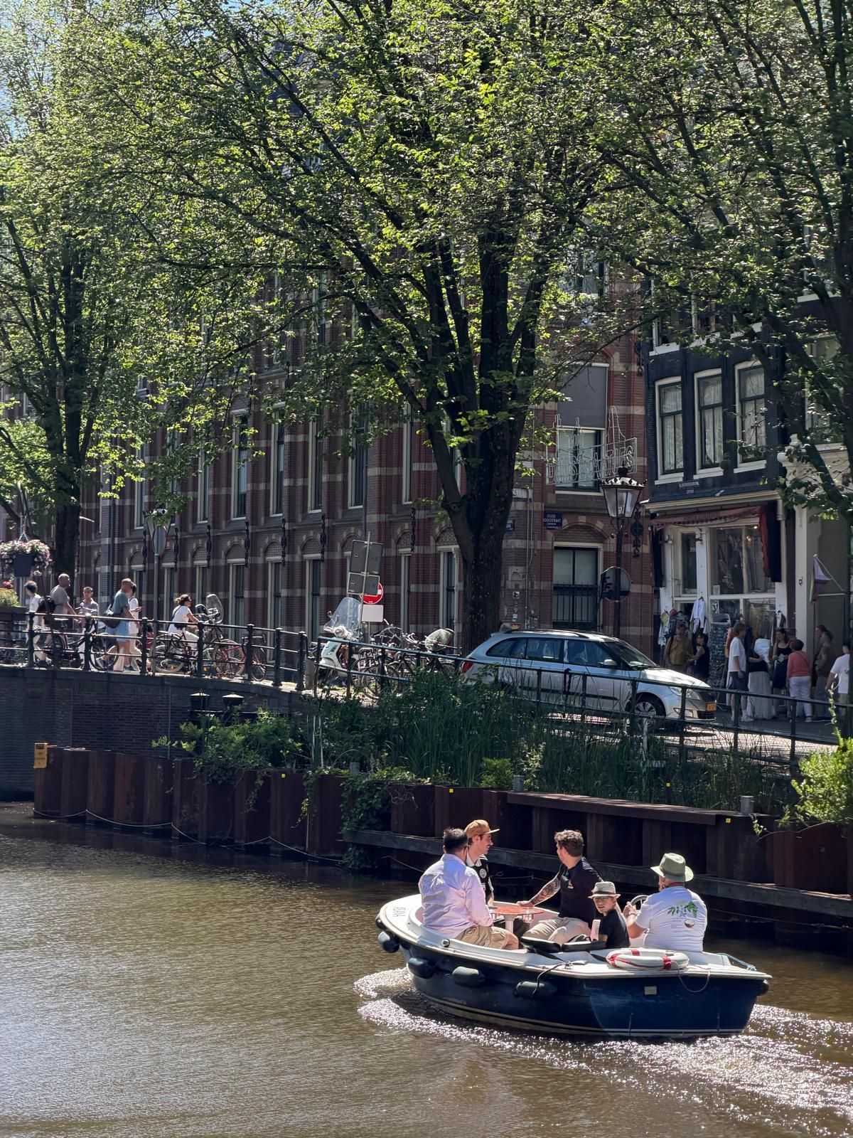 People in a small boat on a canal beside a tree-lined street and brick buildings.