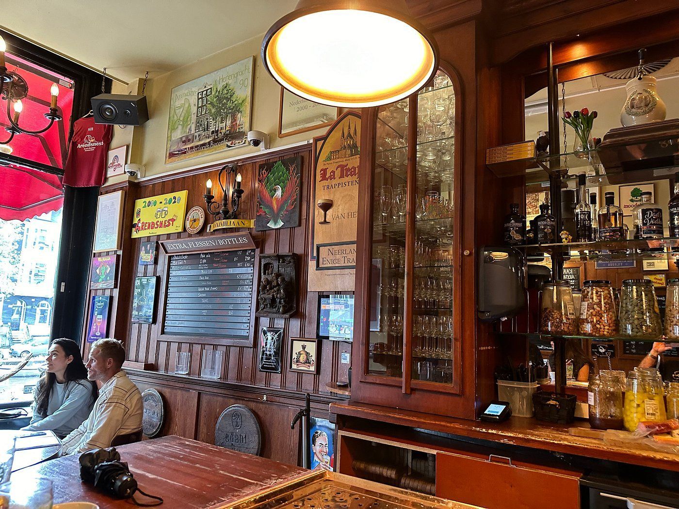 Cozy café interior with mirrored bar, hanging lights, and two seated patrons by the window.