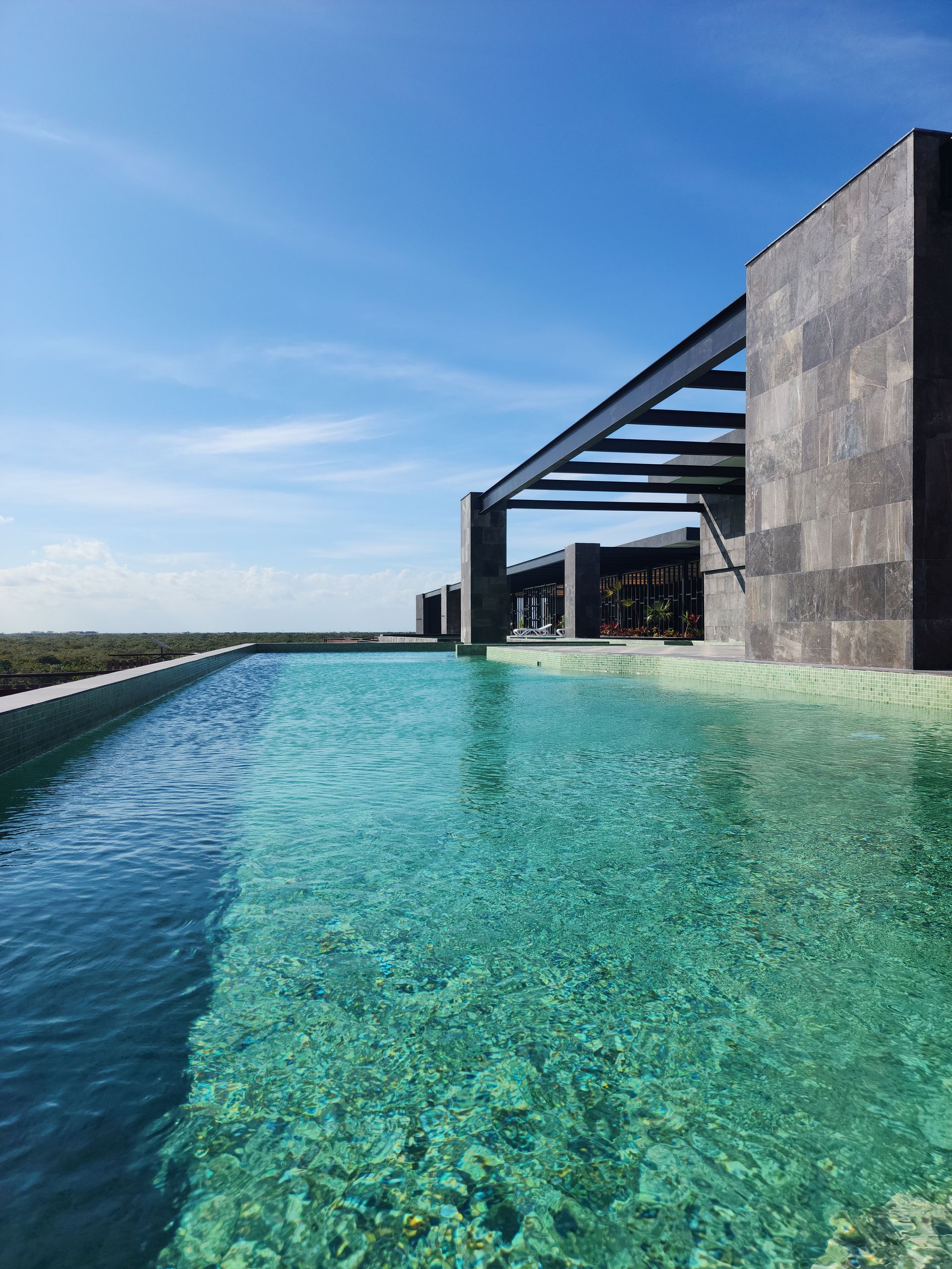 Piscina infinita junto a un edificio de piedra bajo un cielo azul claro, con vistas al océano más allá del borde.