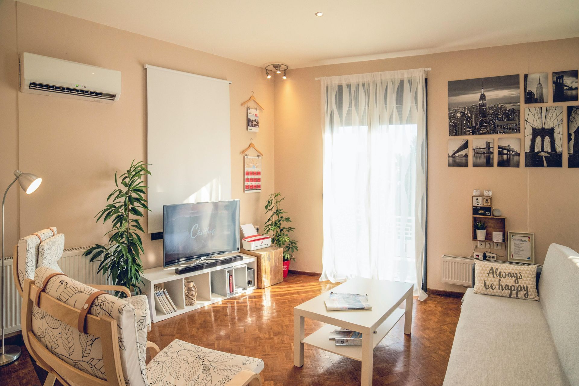 Wohnzimmer mit Fernseher, weißen Möbeln, Pflanzen und einem lichtdurchfluteten Fenster.