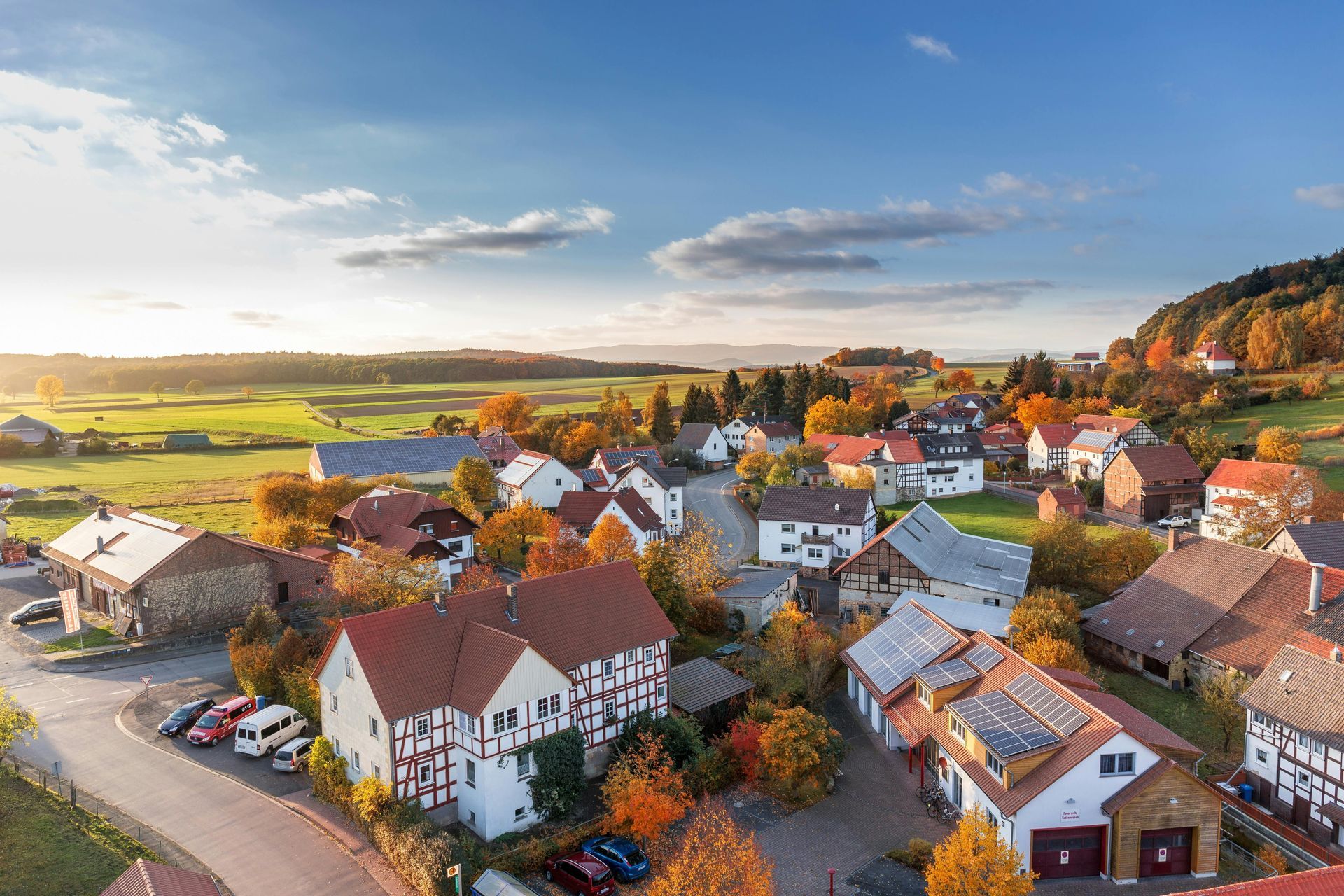 Ein malerisches Dorf mit rotgedeckten Häusern, umgeben von herbstlich gefärbten Bäumen und Feldern unter blauem Himmel.