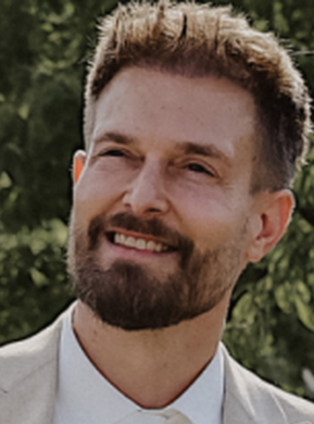 Ein Mann mit kurzem braunem Haar und Bart lächelt; er trägt ein helles Sakko. Die Szene spielt im Freien.