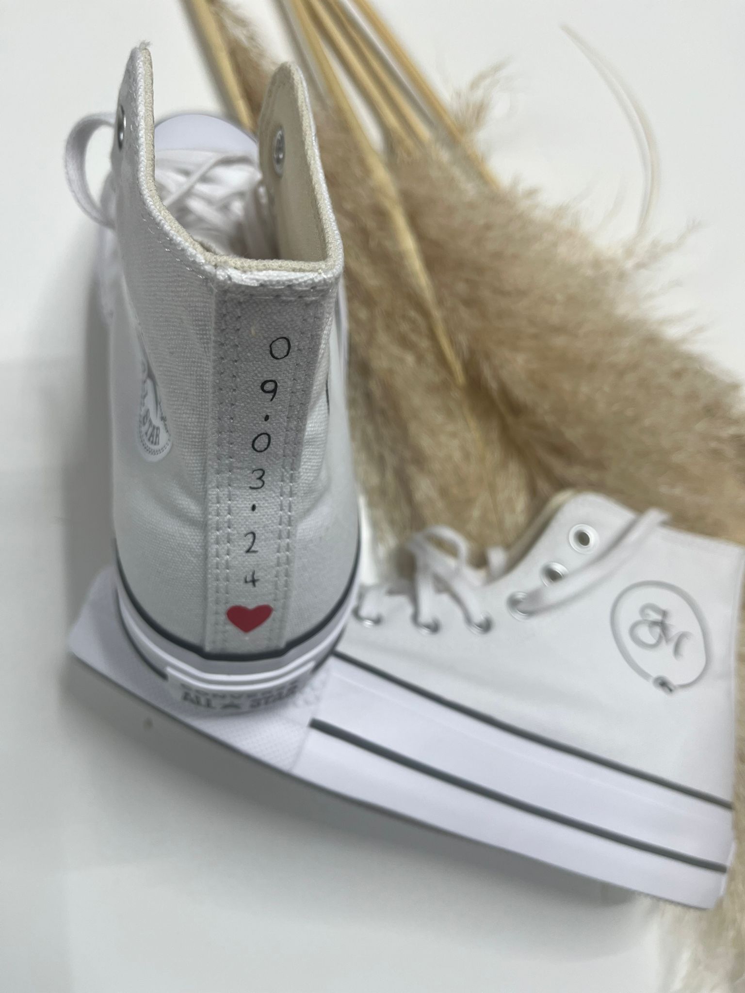 Un par de zapatillas deportivas blancas de caña alta con purpurina, con una fecha y un corazón impresos en la parte trasera, junto a hierba de la pampa seca.