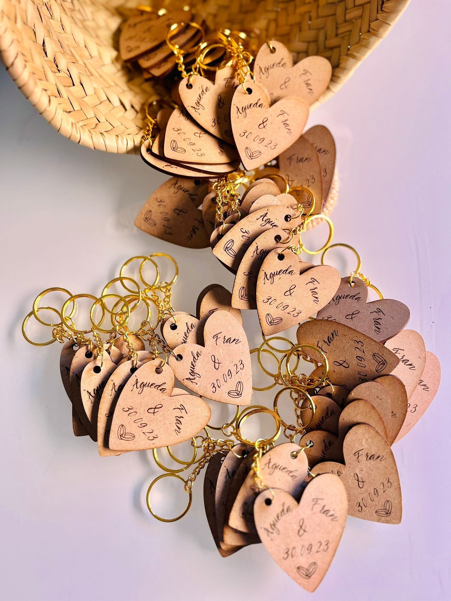 Una cesta repleta de llaveros de madera de color marrón claro con forma de corazón, cada uno grabado con un nombre y una fecha, descansa sobre una superficie blanca.