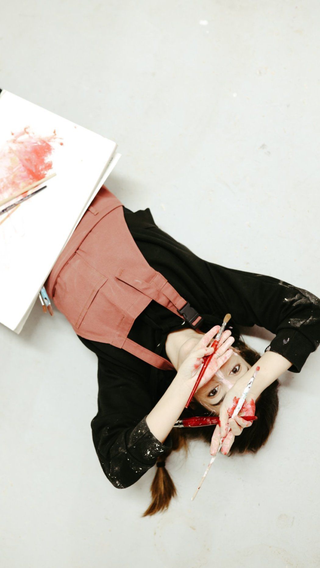 Una artista con un delantal rosa yace en un suelo blanco, sosteniendo pinceles con las manos manchadas de pintura cerca de su rostro.