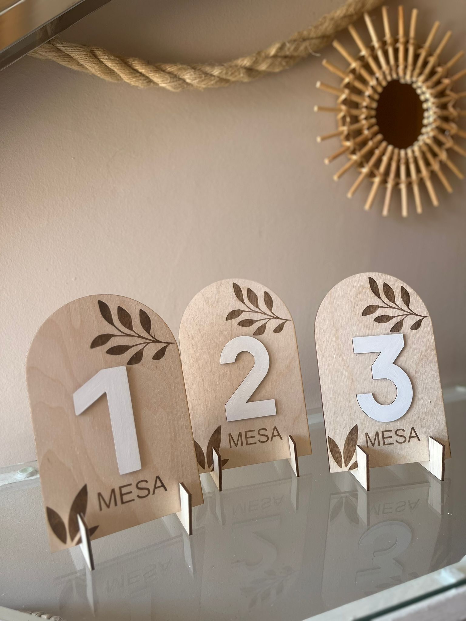 Tres letreros de madera arqueados con los números de mesa 1, 2 y 3, decorados con un diseño de hojas, colocados sobre una superficie de vidrio.