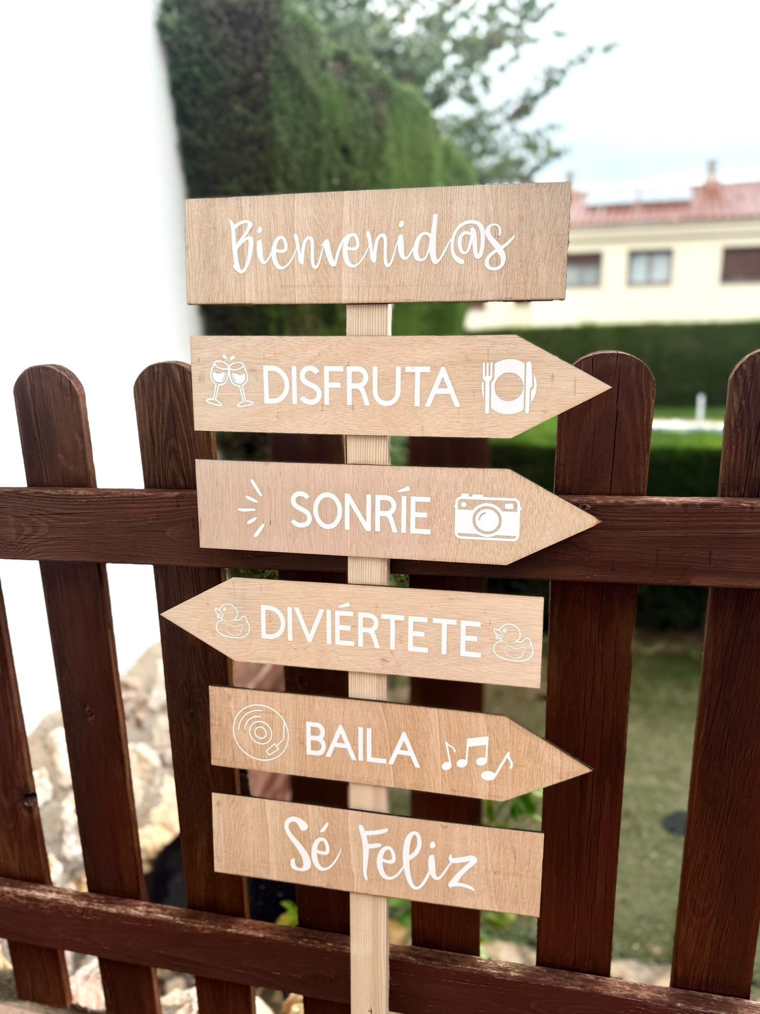 A wooden signpost with six arrows featuring white text in Spanish: Bienvenidos, Disfruta, Sonríe, Diviértete, Baila, Sé Feliz.