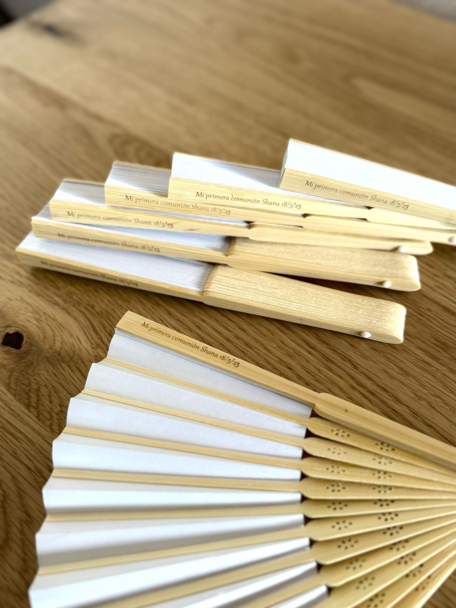 Varios abanicos blancos plegables con armazón de madera clara, uno abierto y los demás plegados, dispuestos sobre una superficie de madera.