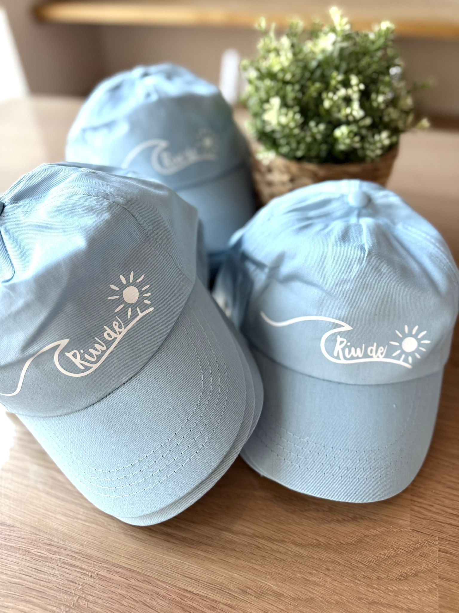 Tres gorras de béisbol azul claro con un diseño de ola y sol bordado en blanco, expuestas sobre una mesa de madera cerca de una planta.