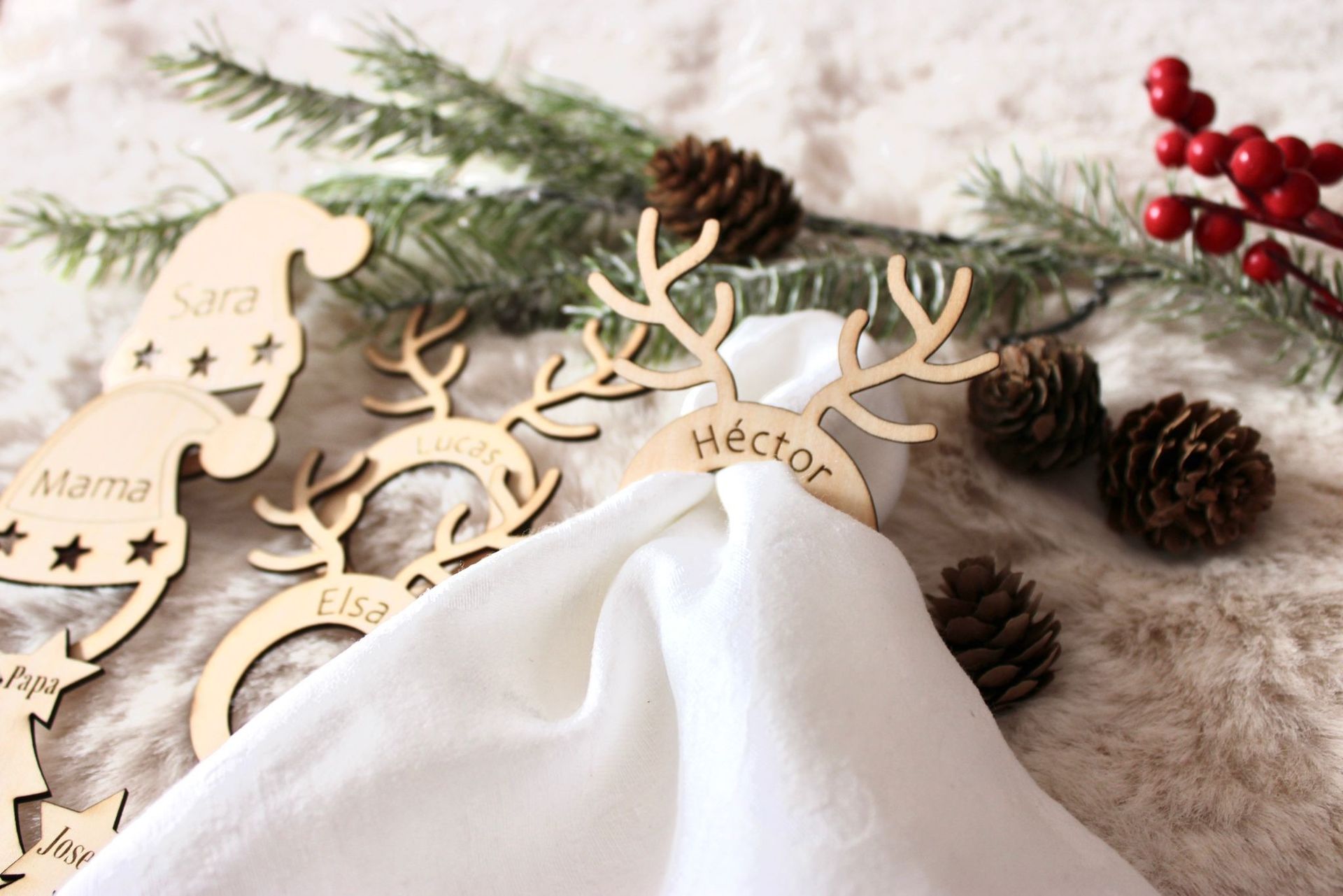 Servilleteros personalizados de madera con forma de reno y gorro de Papá Noel sobre un fondo blanco texturizado con ramas y piñas de pino.