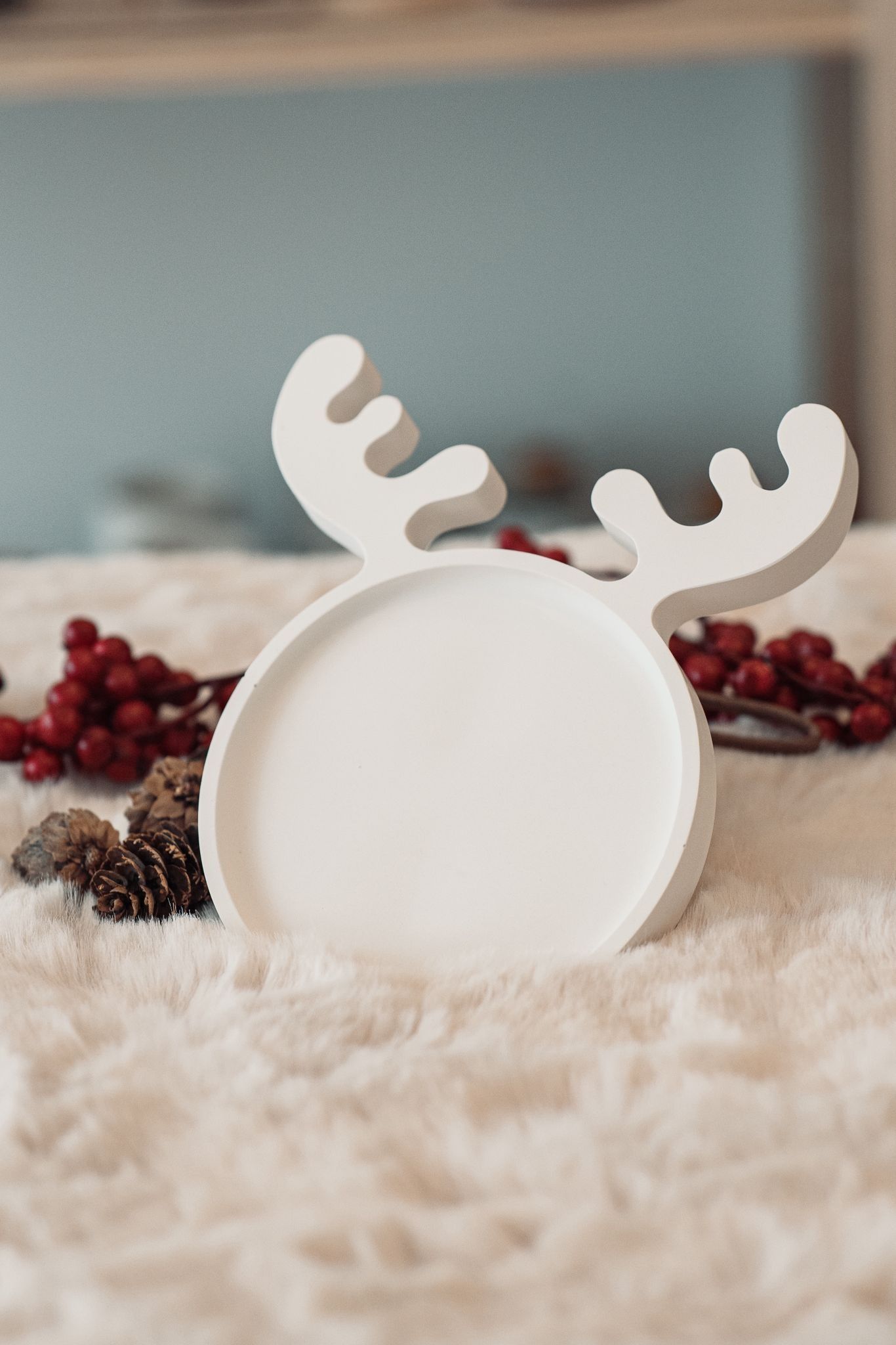Una bandeja decorativa circular blanca con astas de reno, que reposa sobre una superficie suave y texturizada con piñas y bayas rojas.