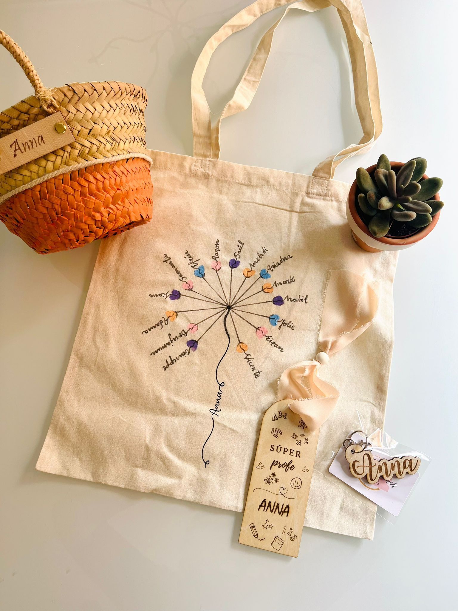 Un bolso de lona con un diseño de diente de león bordado, una cesta tejida, una pequeña planta suculenta en maceta y una etiqueta de madera.