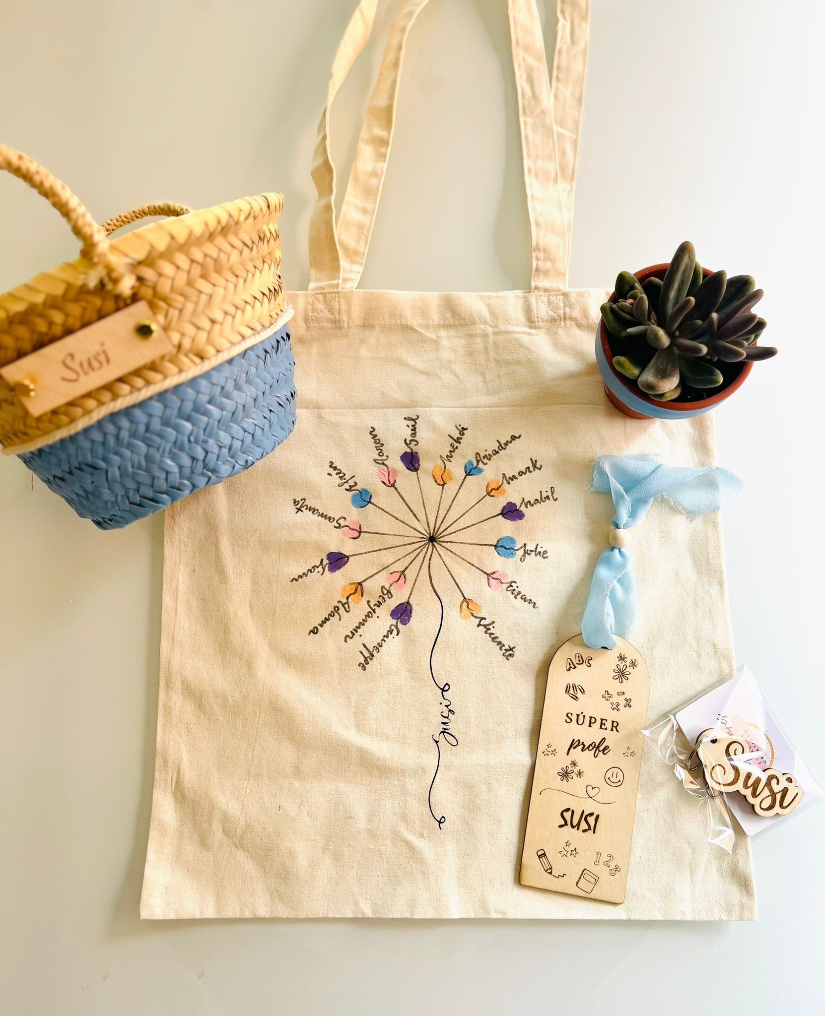 Una cesta de paja, una bolsa de tela con motivos florales, un marcapáginas y una pequeña planta suculenta dispuestos sobre una superficie clara.