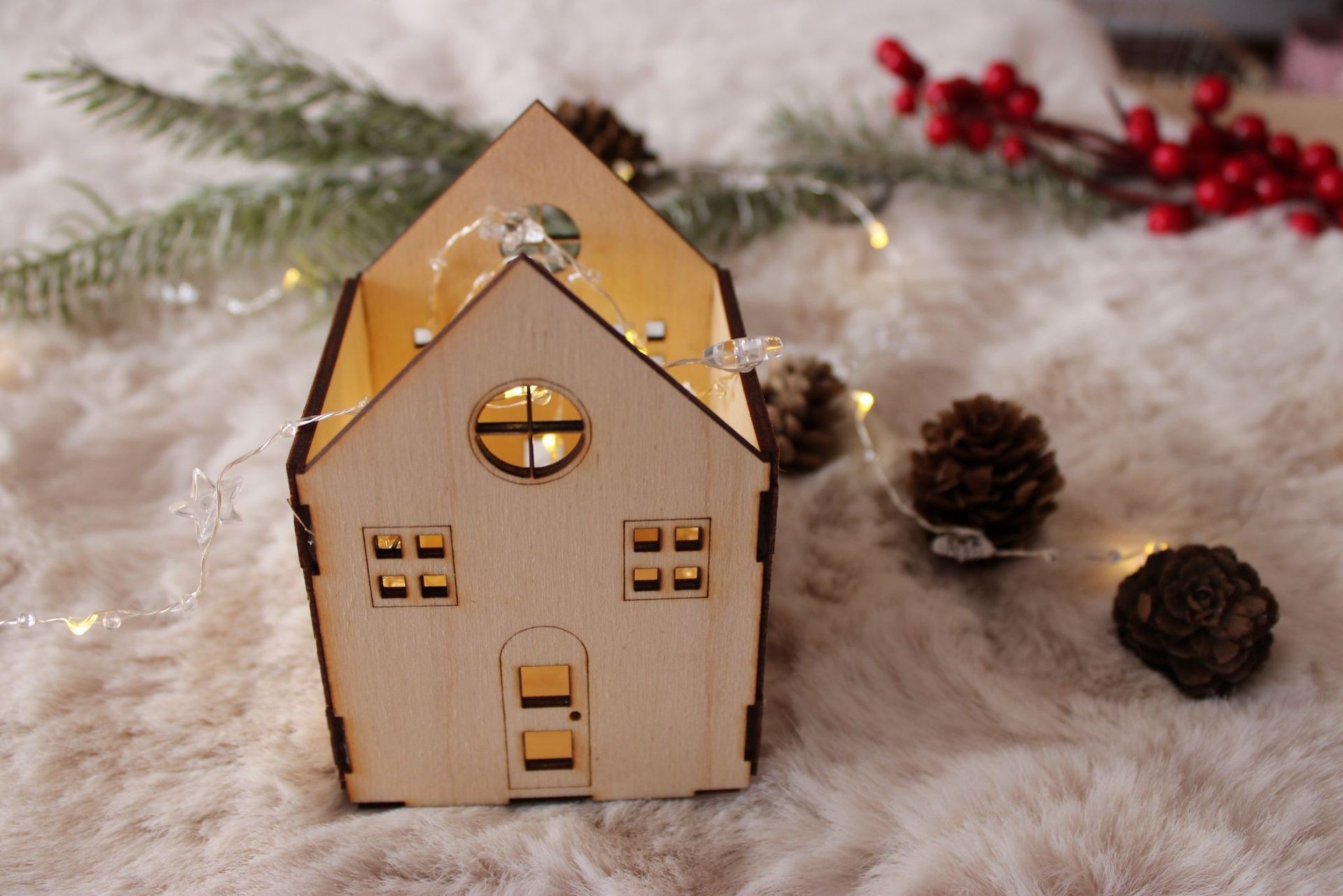 Un adorno de madera con forma de casa que se ilumina, sobre una superficie blanca y esponjosa, decorado con piñas, bayas de acebo y luces de hadas.