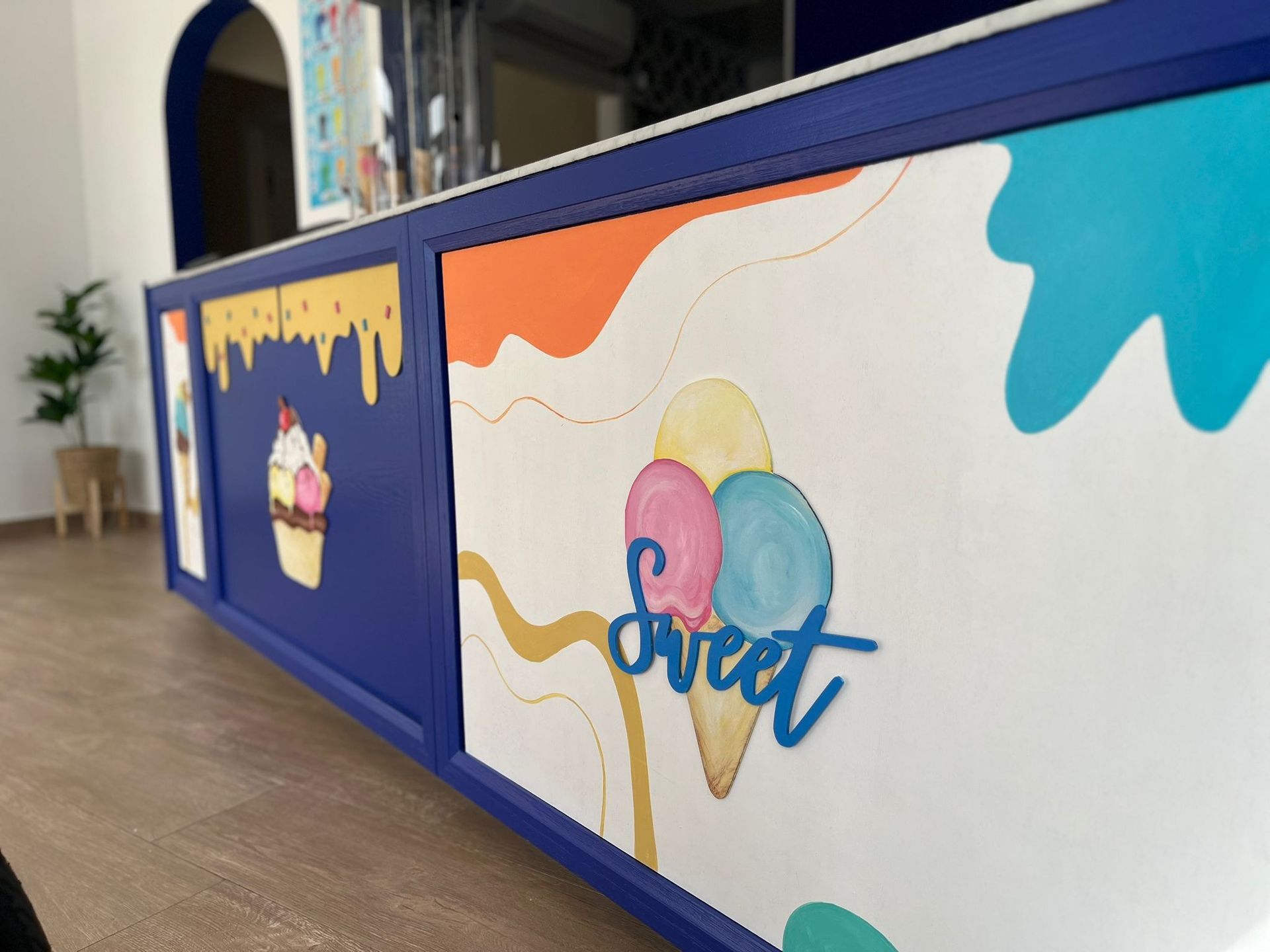 Un mostrador de heladería vibrante, con marco azul, murales coloridos, bolas de helado pintadas y un letrero azul que dice 