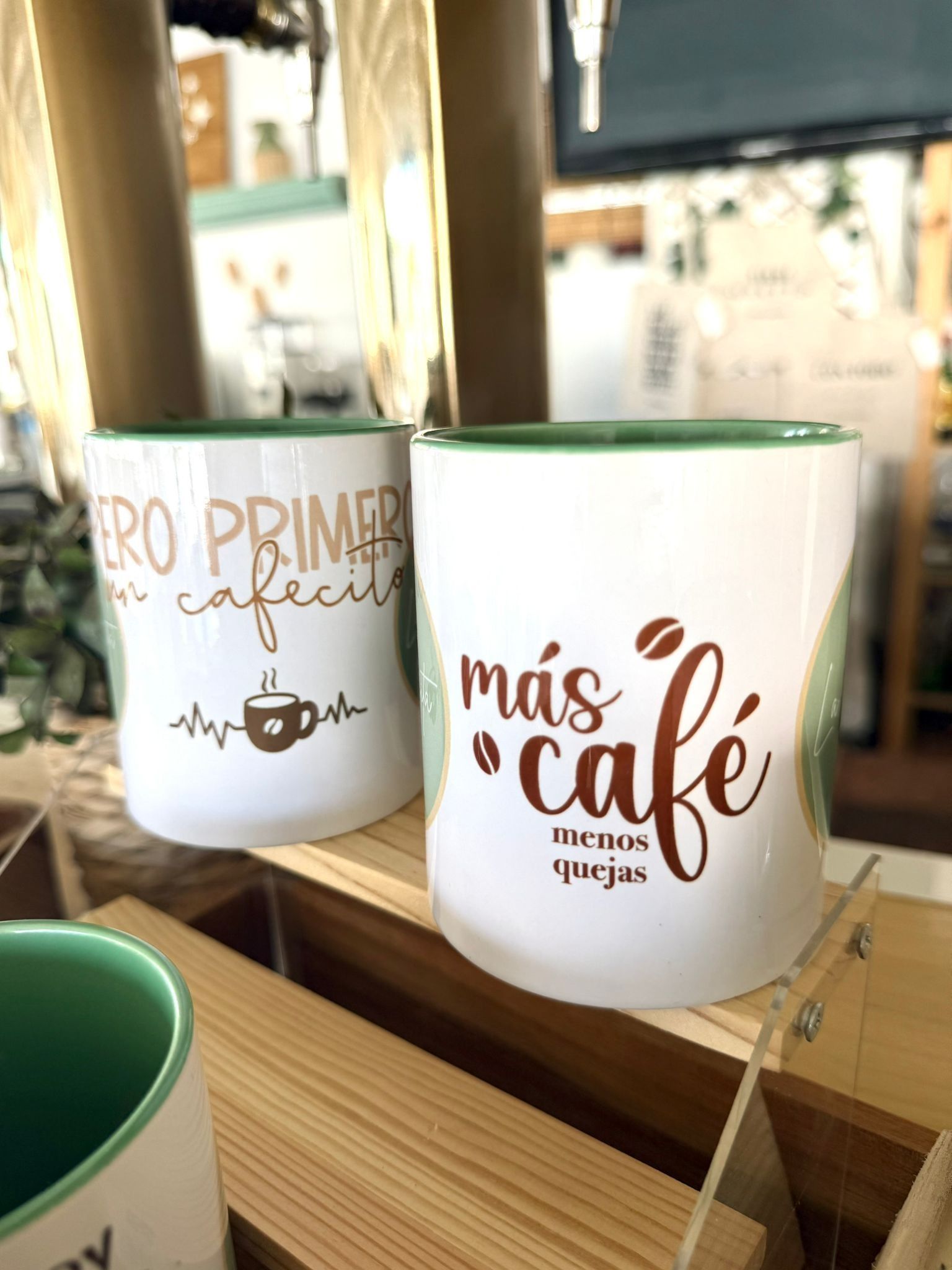 Dos tazas de cerámica blanca con interior verde y texto en español con motivos relacionados con el café reposan sobre una estantería de madera.