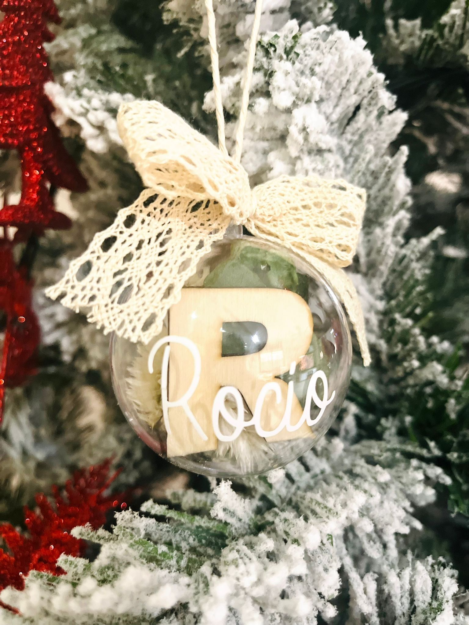 Un adorno navideño de cristal transparente colgado de un pino nevado, con la letra R y el nombre 