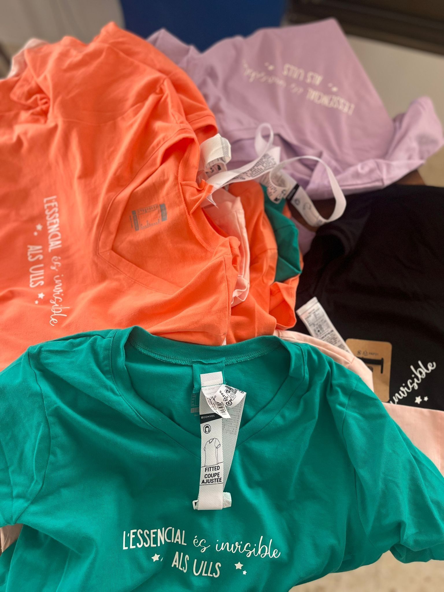 Una pila de cuatro camisetas dobladas de color naranja, morado, verde y negro, cada una impresa con el texto 