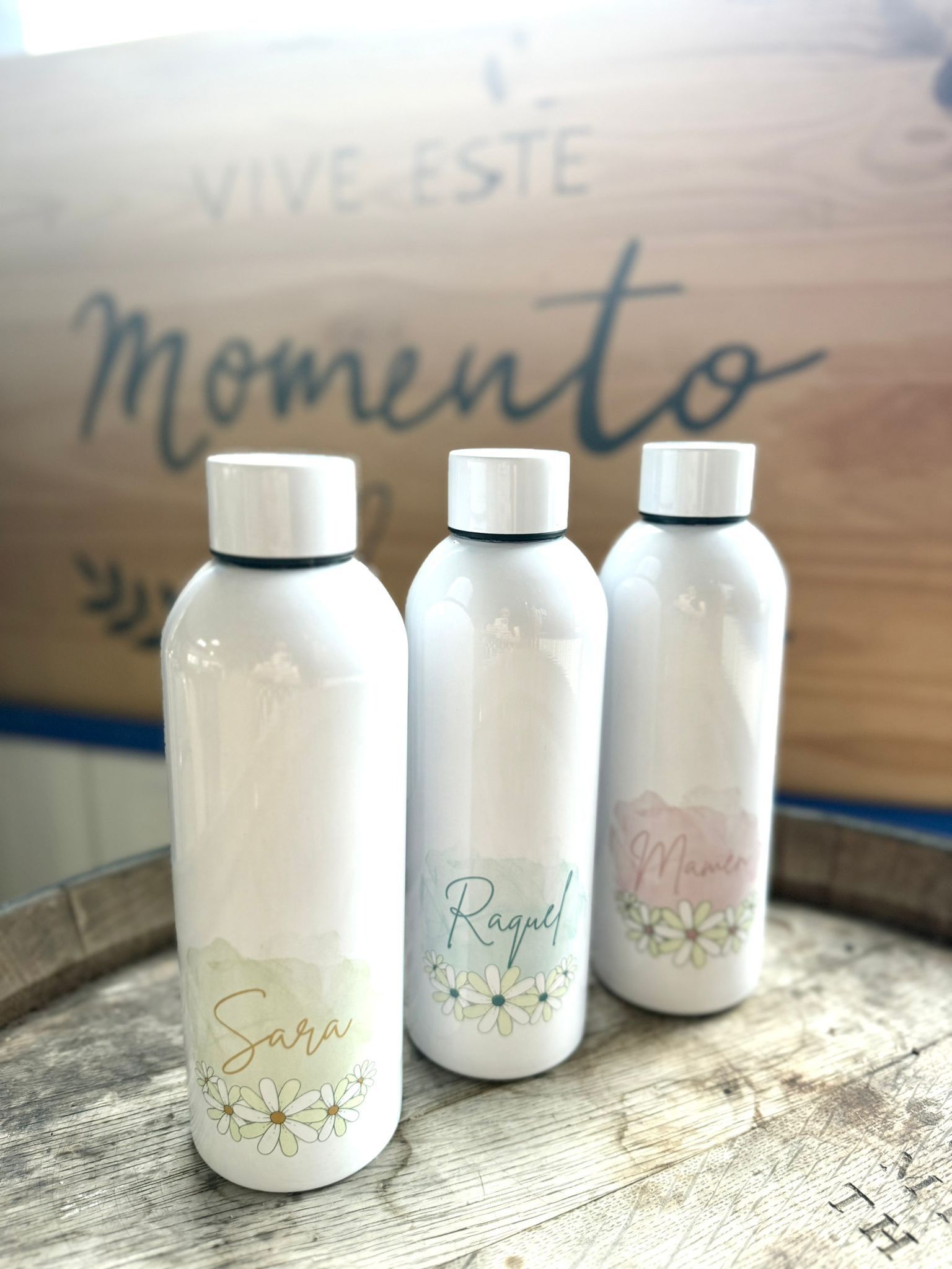 Tres botellas de agua blancas personalizadas con diseños florales, colocadas sobre un barril de madera frente a un letrero que dice 