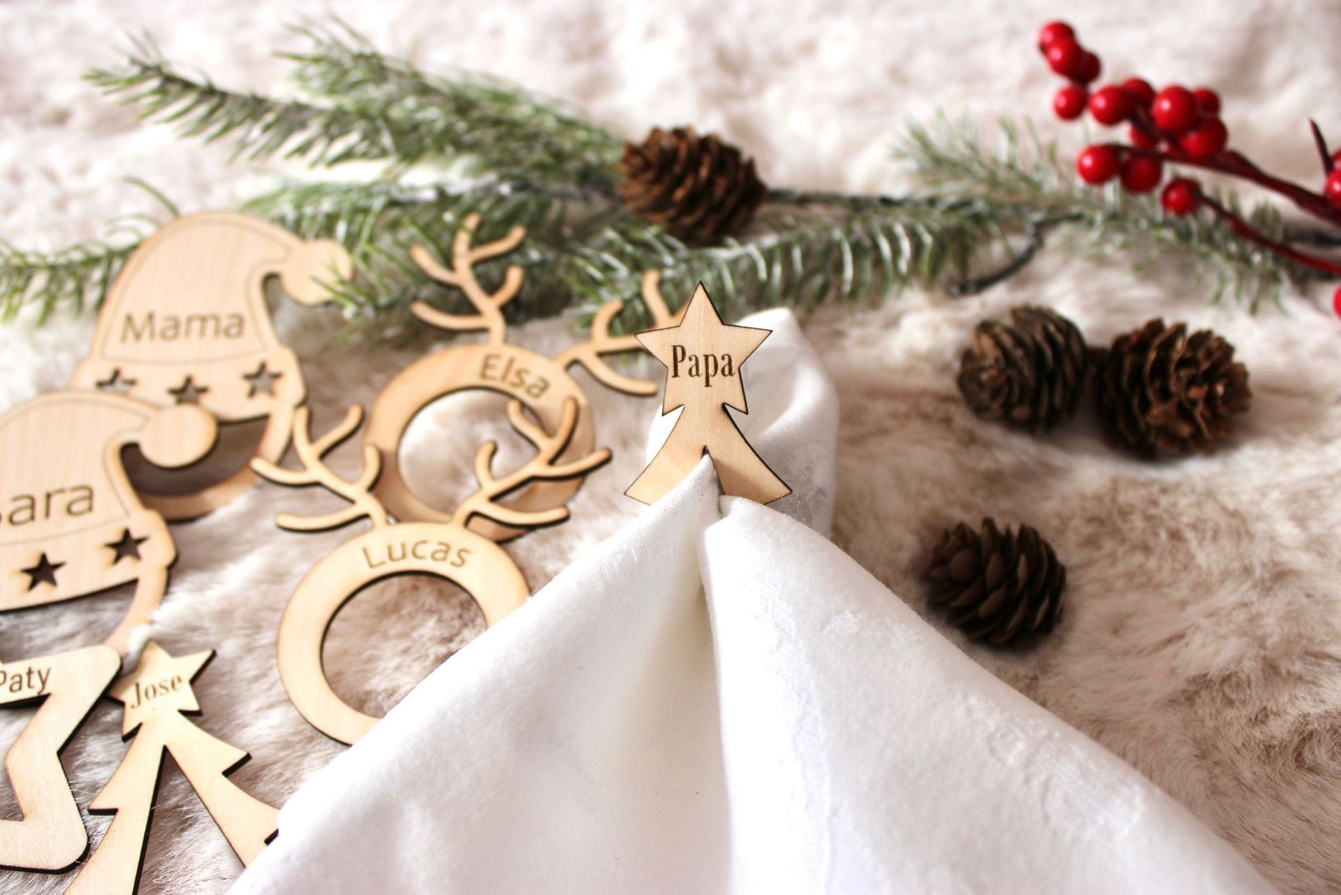 Anillos para servilletas de madera navideños personalizados sobre una servilleta de tela blanca, rodeados de ramas de pino, piñas y bayas de acebo.