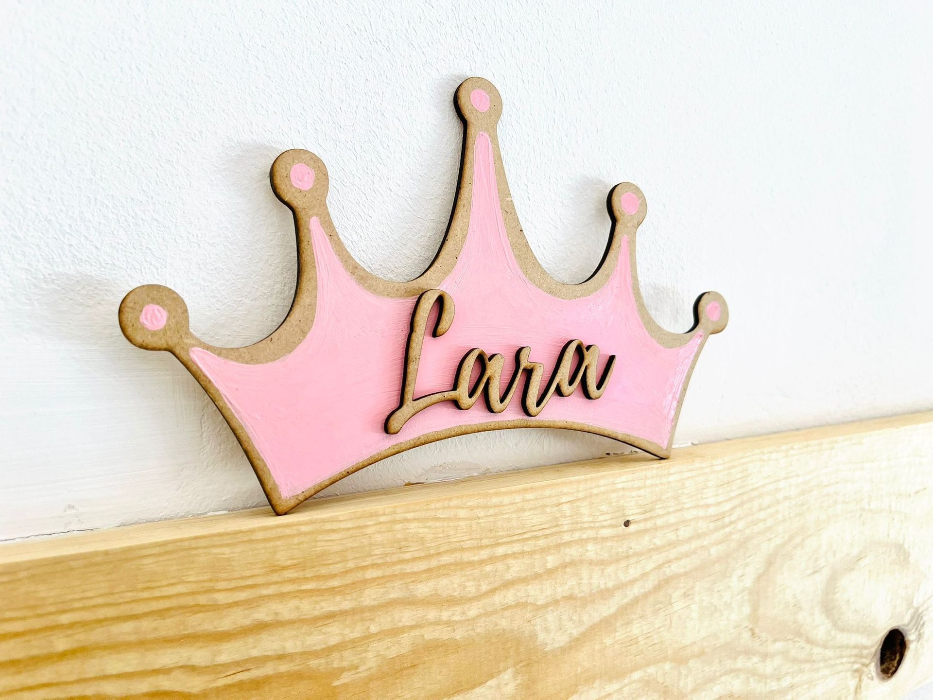 Un adorno de pared en forma de corona de madera rosa con el nombre 