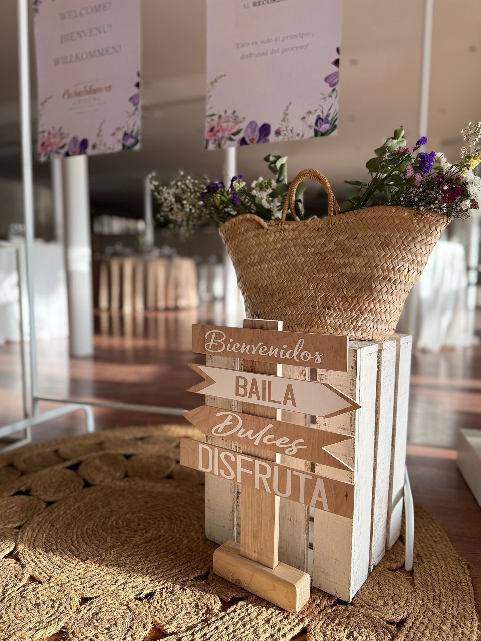 Una cesta de paja con flores reposa sobre una caja de madera blanca con letreros que dicen 