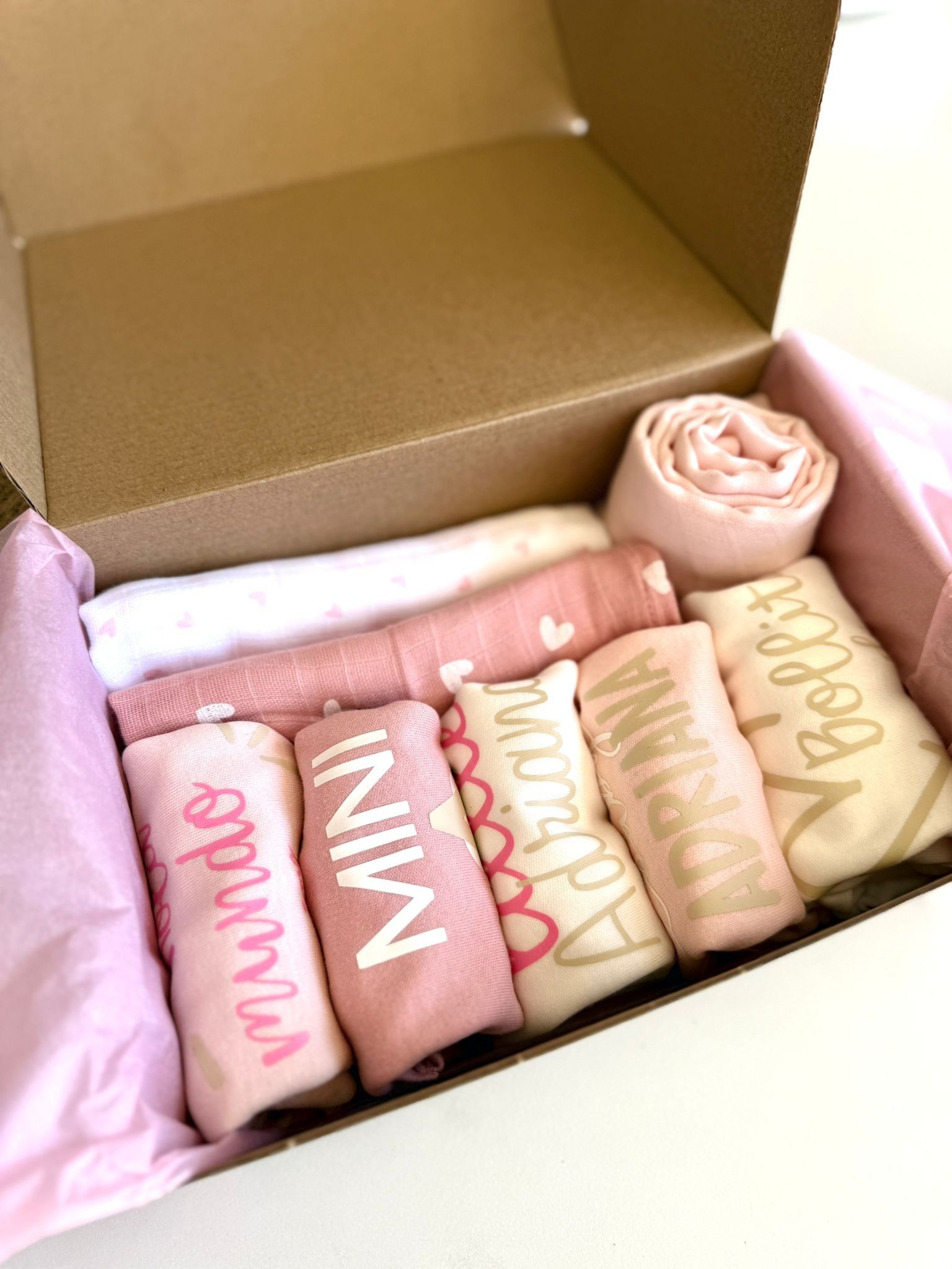 Una caja de cartón abierta, forrada con papel de seda rosa, llena de ropa de bebé doblada y personalizada en tonos rosas.