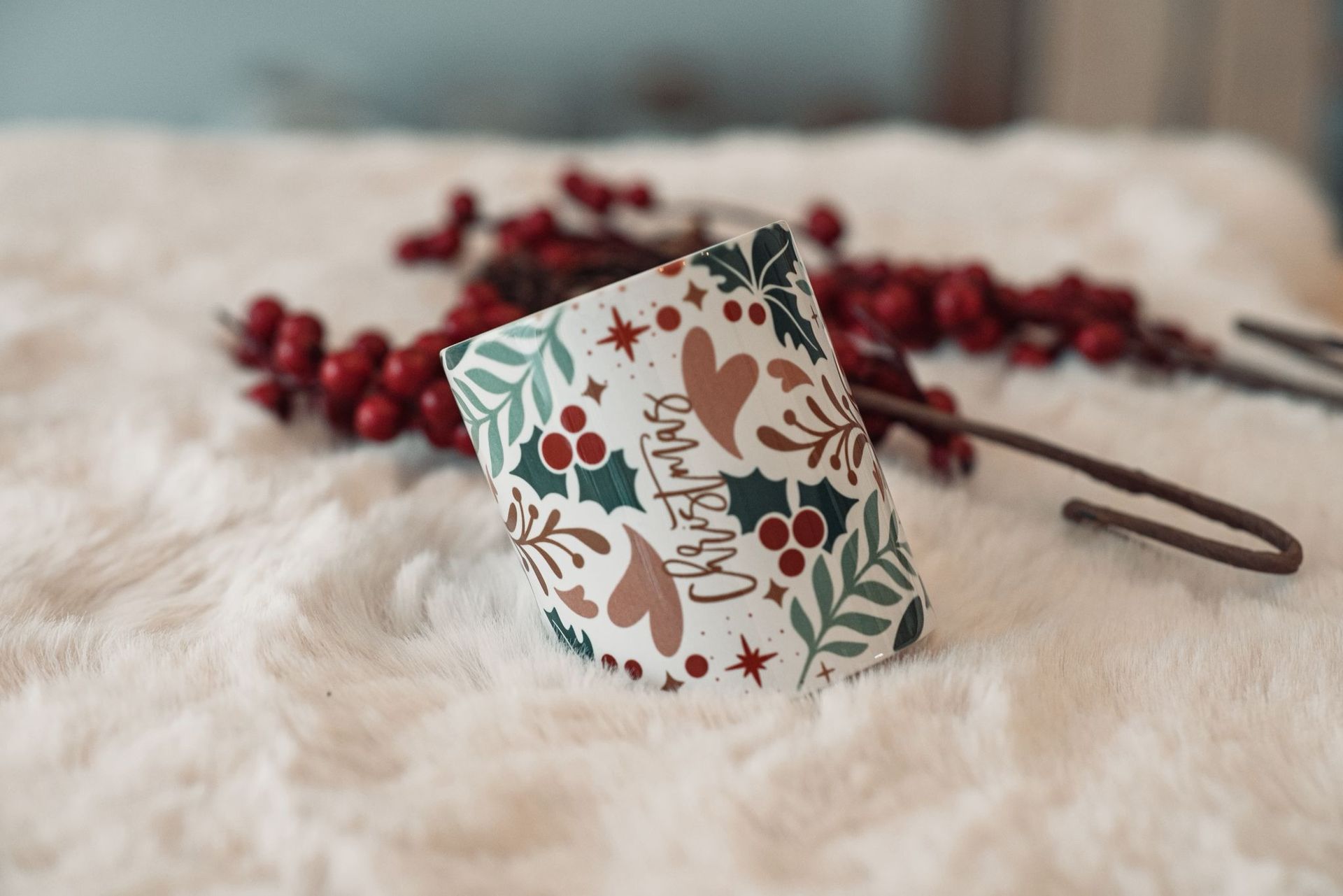 Una taza con motivos navideños reposa sobre una superficie de piel blanca, rodeada de bayas rojas de acebo y un gancho de metal, con un diseño festivo navideño.