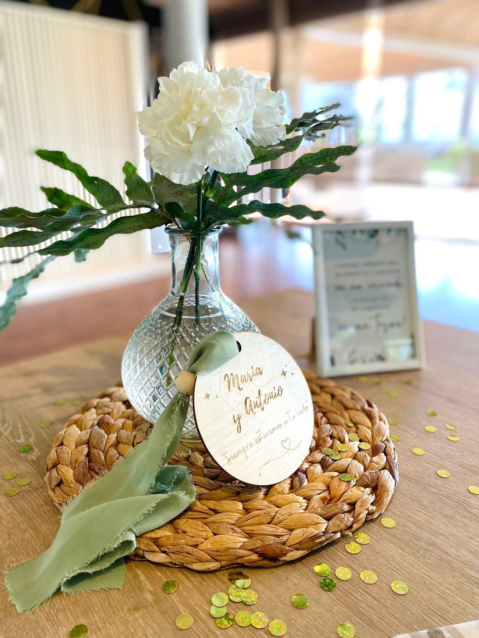Una flor blanca en un jarrón de cristal sobre una estera tejida, con una etiqueta de madera personalizada y confeti sobre una mesa de madera.