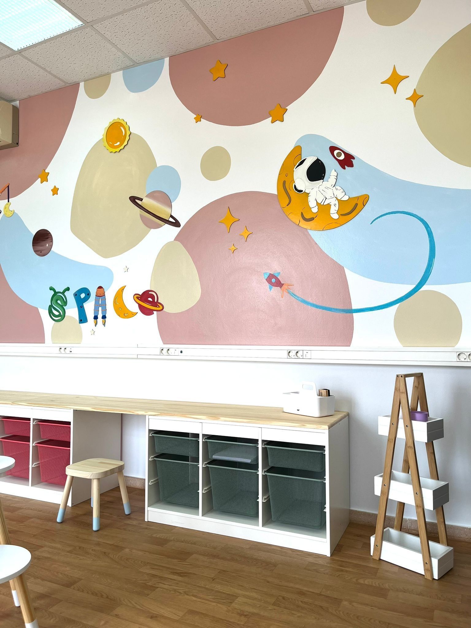 Un colorido mural de pared con temática espacial que representa a un astronauta de dibujos animados sobre una luna creciente, situado encima de los muebles de almacenamiento del aula.