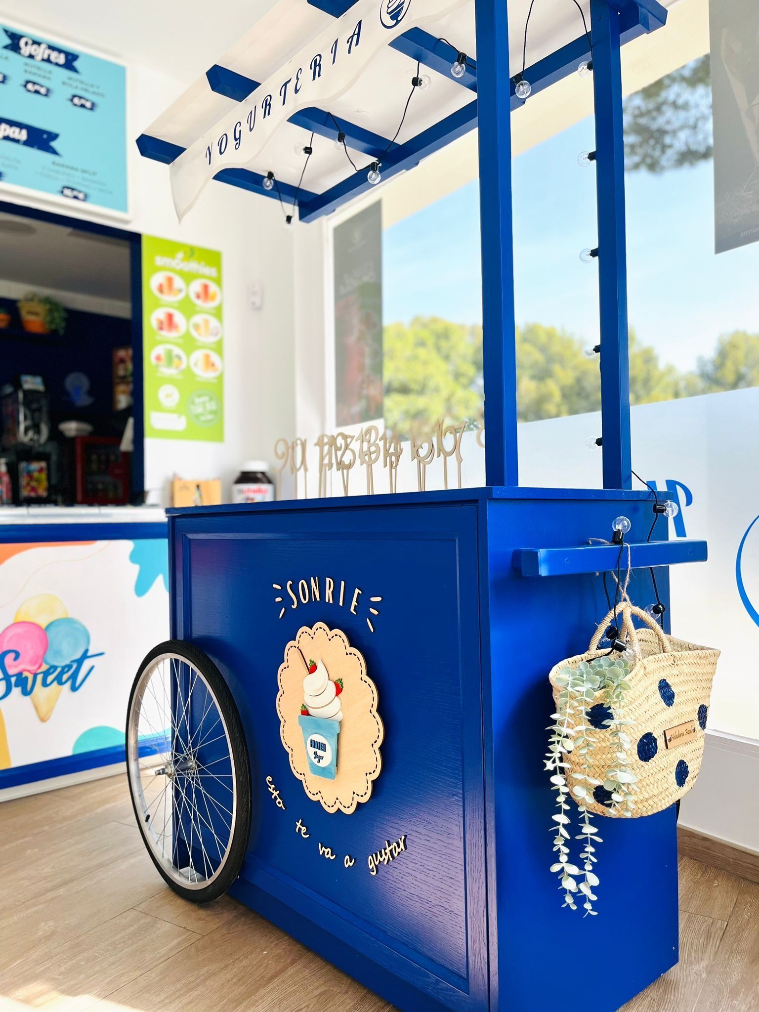 Un carrito de helados de color azul brillante se encuentra en una zona al aire libre, con una bolsa de paja colgando de un lado y una pizarra con el menú cerca.