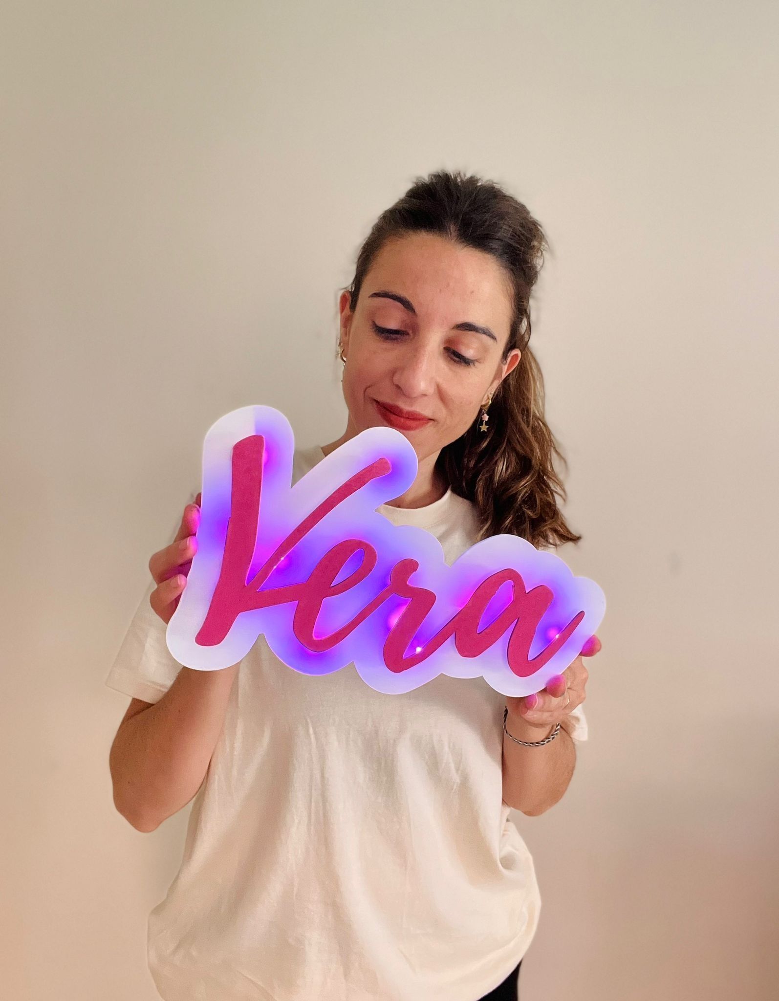 Una persona con cabello rizado sonríe mientras sostiene un letrero de neón rosa brillante que dice 